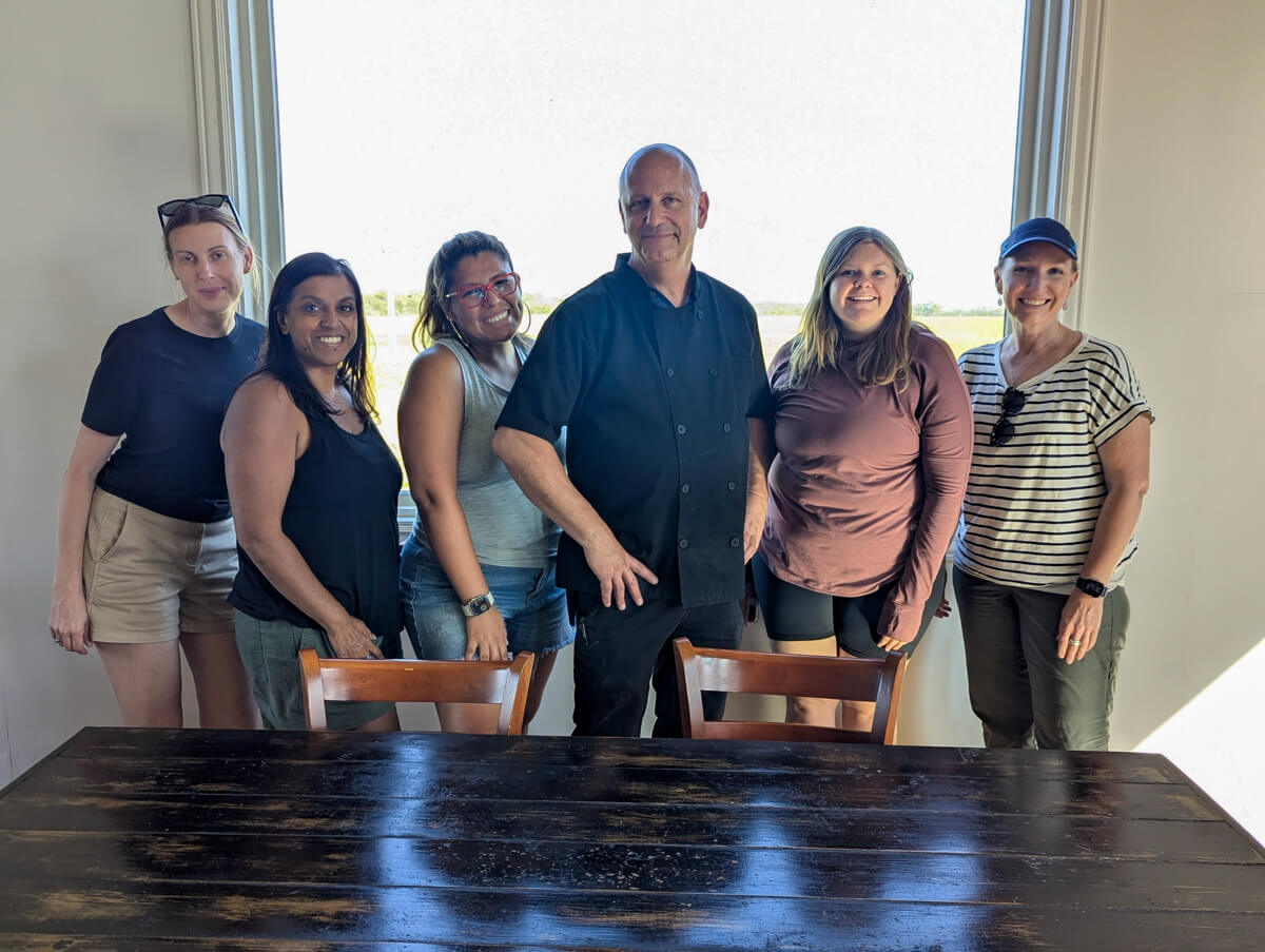 A group of 6 in front of a window with a wooden table in front. Chef Stan in the middle.