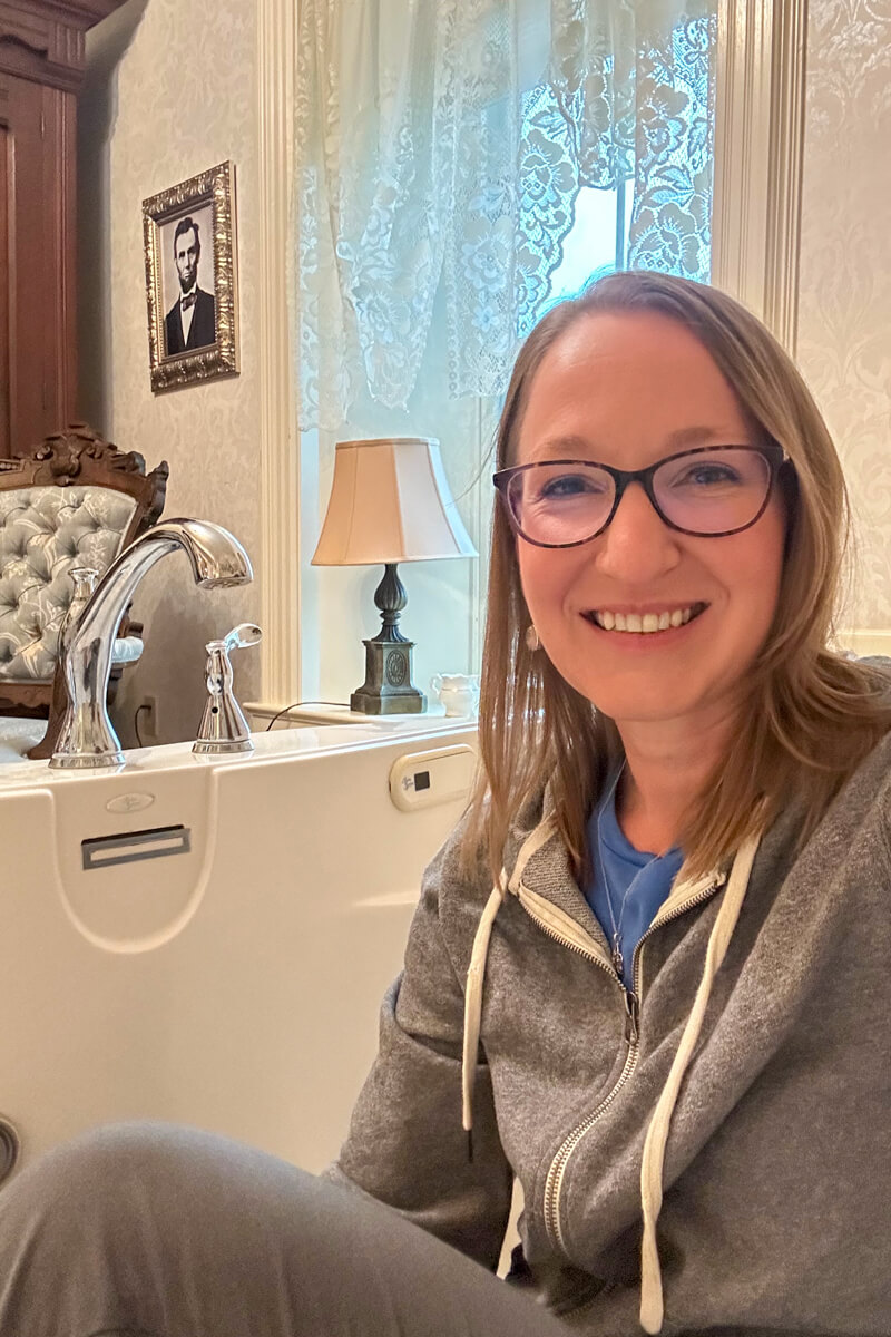 A blond woman in glasses and a grey zip hoodie sitting in a giant white bathtub with a picture of Abraham Lincoln on the wall behind her
