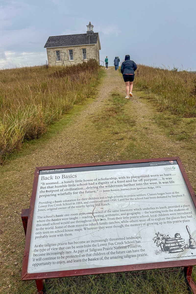 A white sign in front of a pathway with people walking up to the one room school house