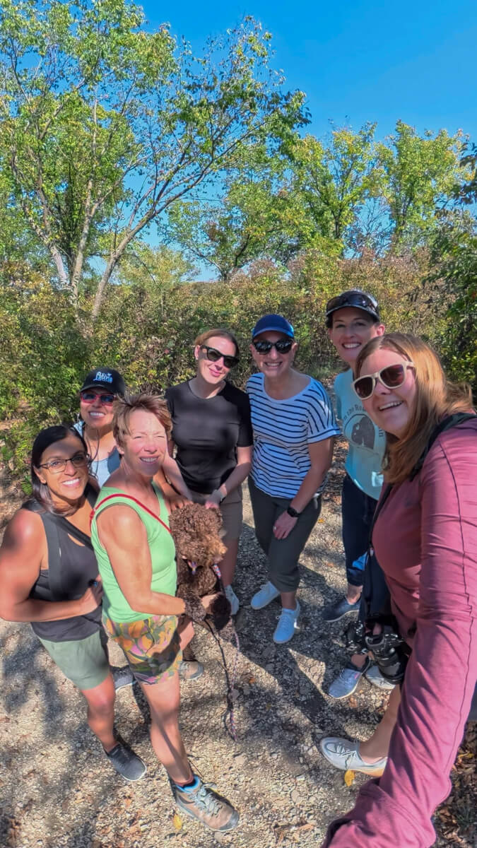 A group of 7 women and a brown dog with bushes and trees in the background