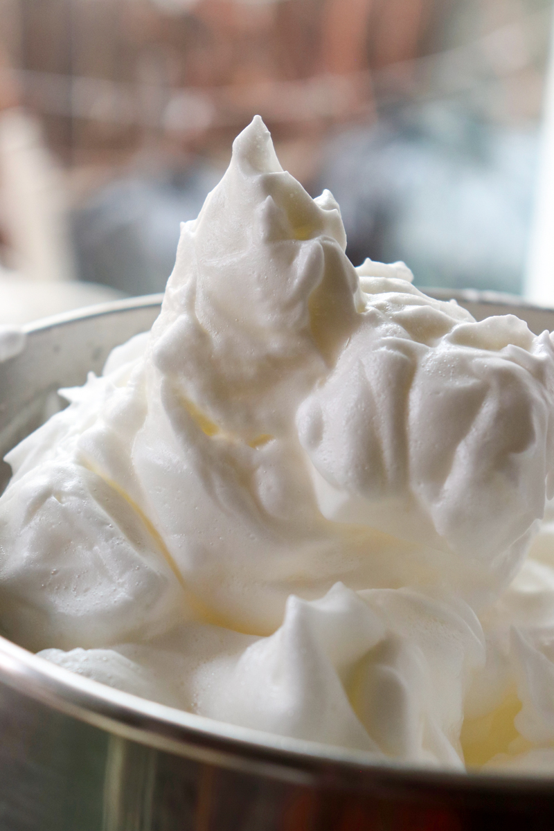 Close up side view of a metal bowl with a spatula stirring the whipped egg whites