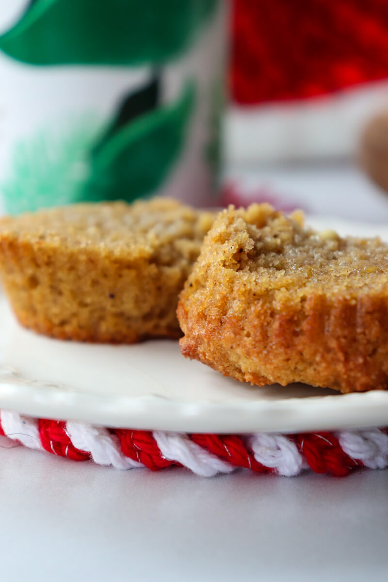 A low carb gingerbread muffin cut in half on a white plate with melted butter in front of a white mug with green leaves on it