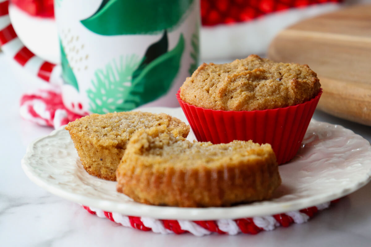 A white plate with two muffins, one still in a red silicone cup and the other cut in two halves with melted butter. A white mug with green leaves is in the background.