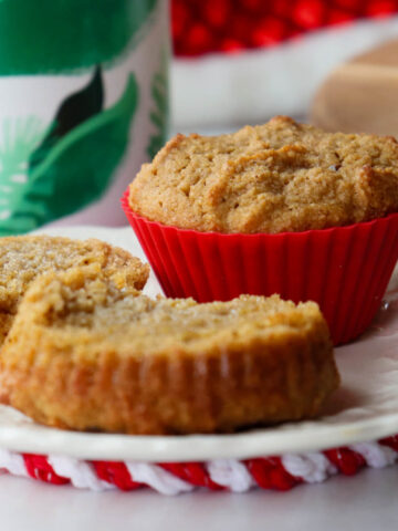 A white plate with two muffins, one still in a red silicone cup and the other cut in two halves with melted butter. A white mug with green leaves is in the background.
