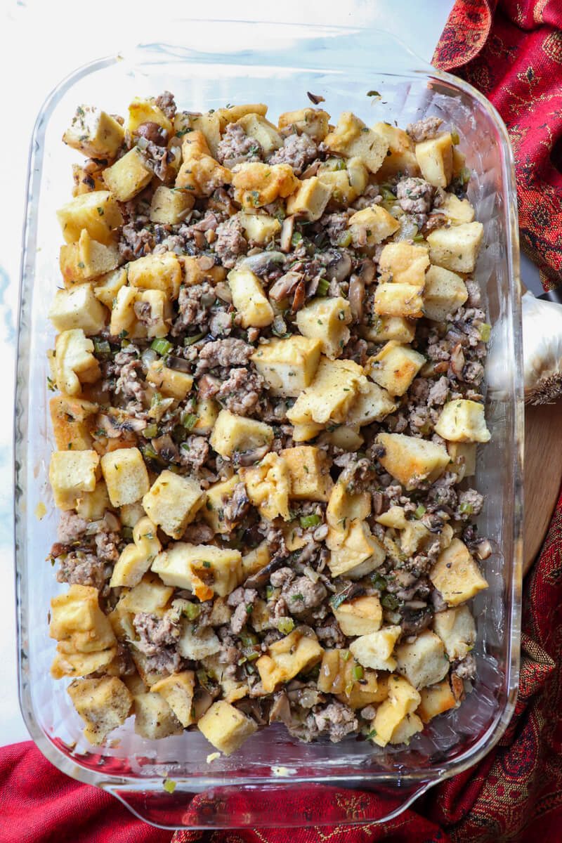 Overhead view of glass dish of uncooked keto stuffing with toasted protein bread and sausage on a circular board with a red cloth next to it