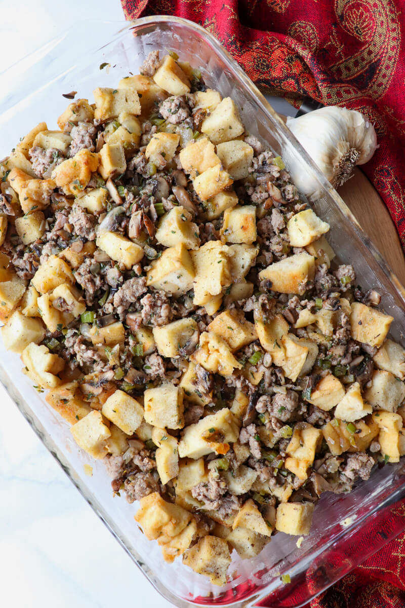 Overhead angled view of glass dish of uncooked keto stuffing with toasted protein bread and sausage on a circular board with a red cloth next to it