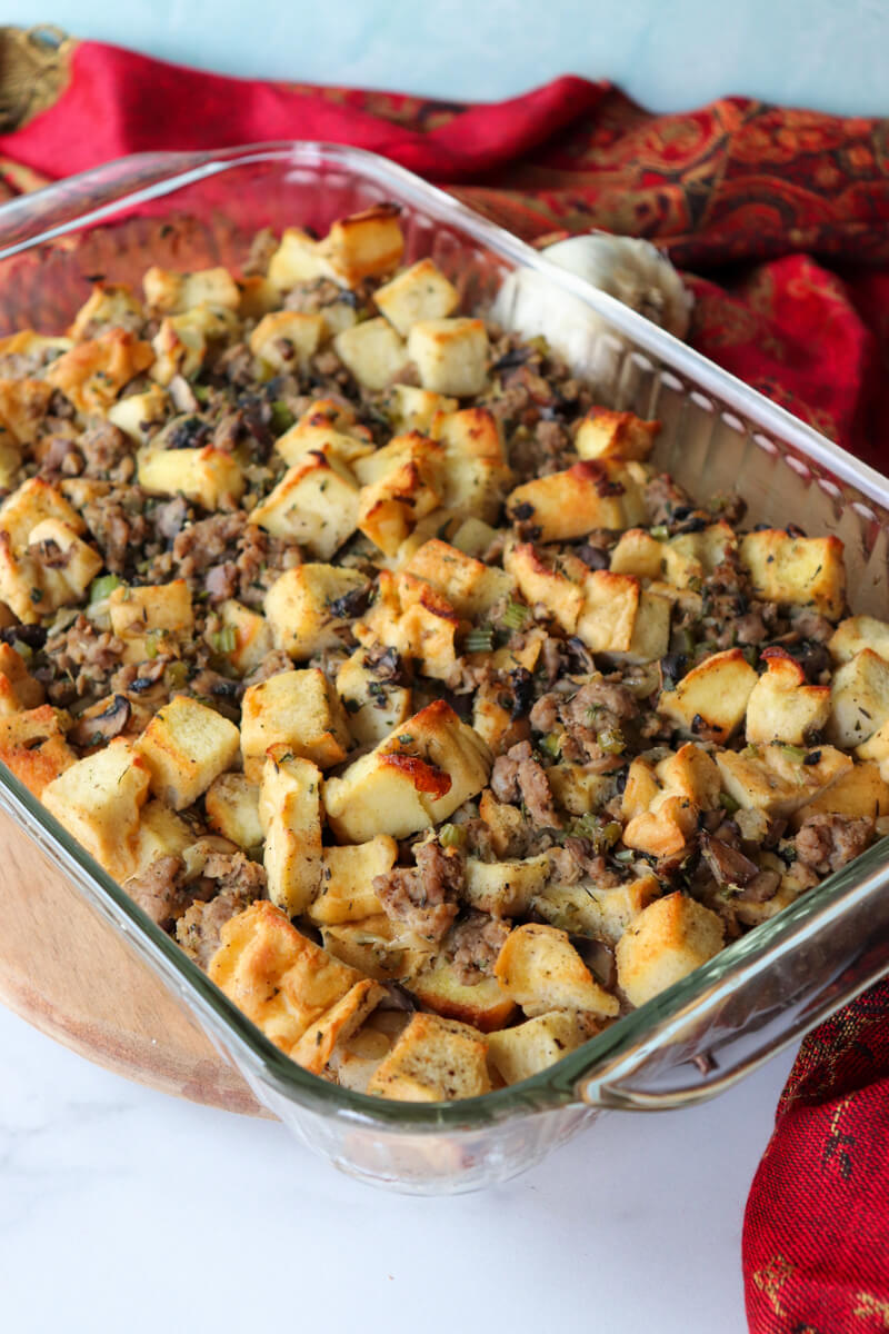 A glass dish of cooked keto stuffing with toasted protein bread and sausage on a circular board with a red cloth next to it