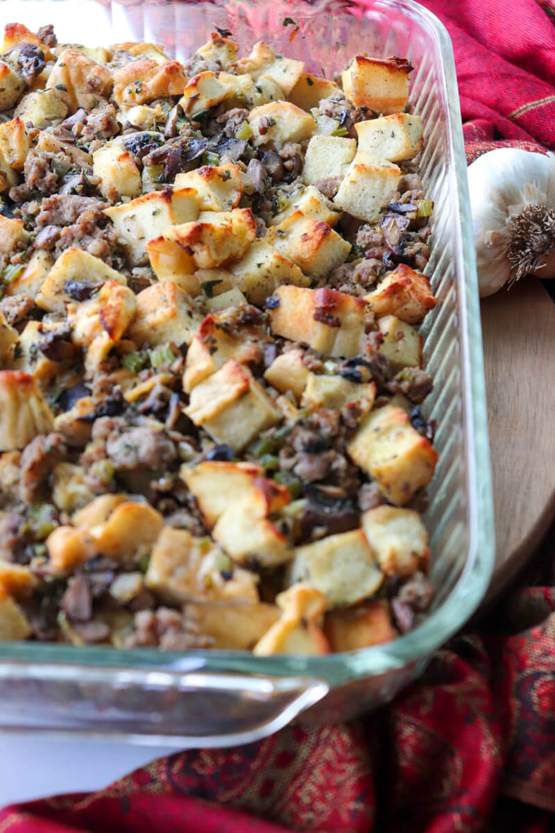 Overhead view of a glass dish of cooked keto stuffing with toasted protein bread and sausage on a circular board with a red cloth next to it