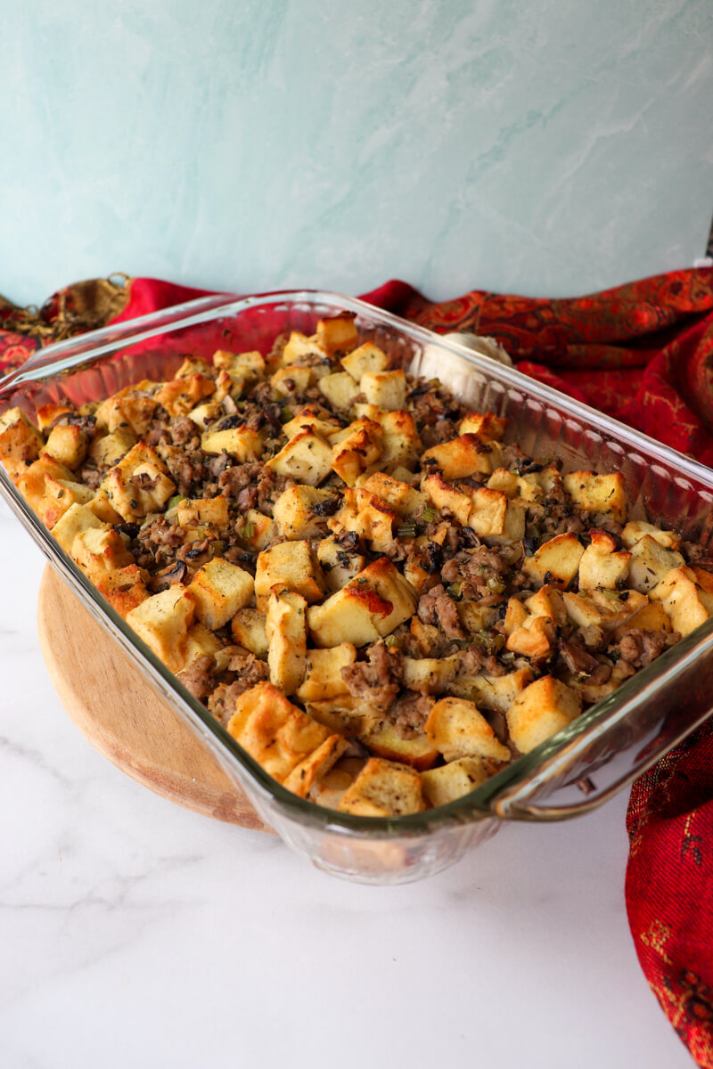 A glass dish of cooked keto stuffing with toasted protein bread and sausage on a circular board with a red cloth next to it