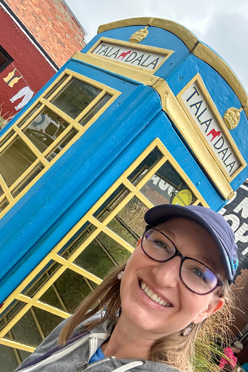 A blue and yellow phone booth that says Tala Dala on the top with a blond woman in a navy baseball cap and glasses in front