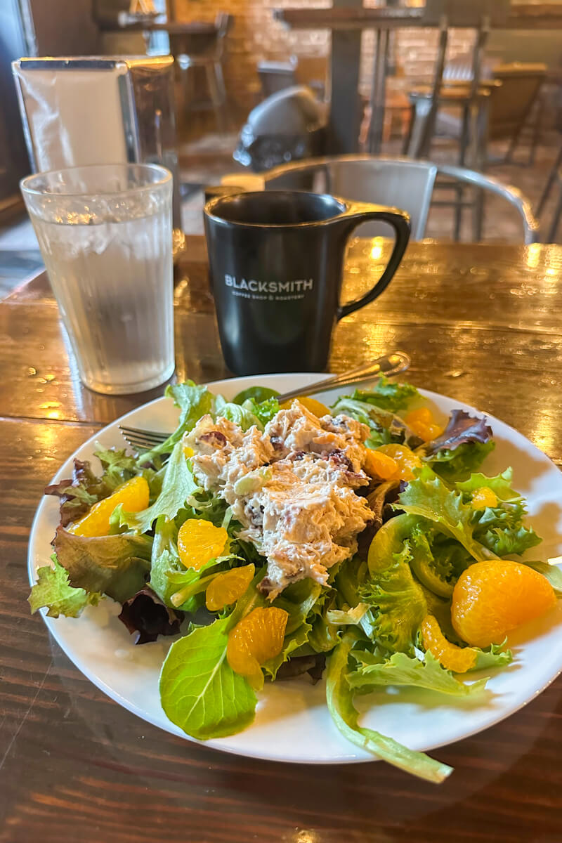 A white plate of chicken salad with mandarins in front of a black coffee cup on the wooden table