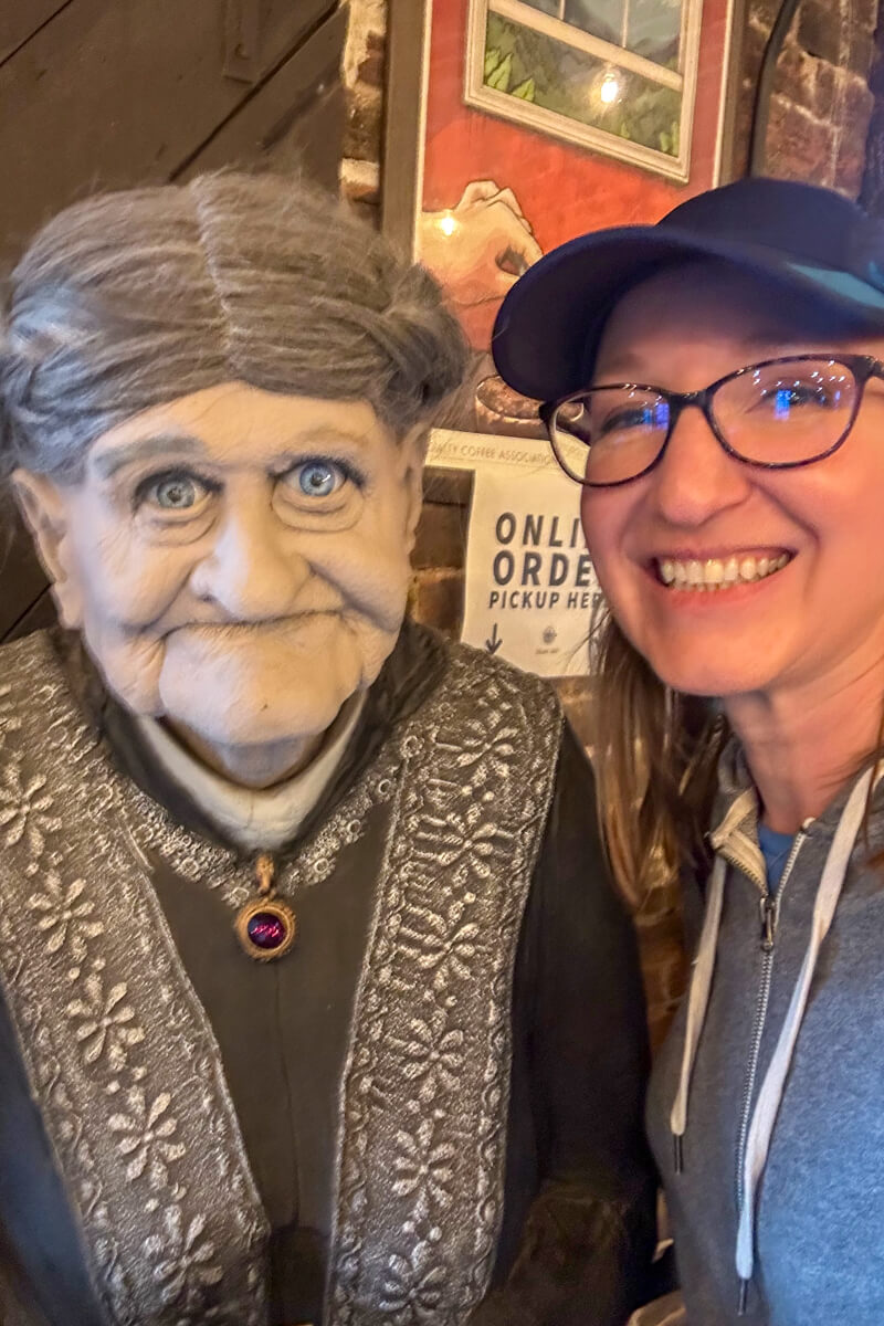 A blond woman with glasses and a navy cap posing with a gray haired old woman mannequin
