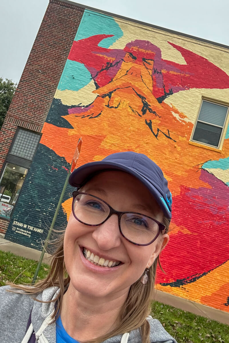 A blond woman in glasses and navy cap in front of an orange and red Viking mural