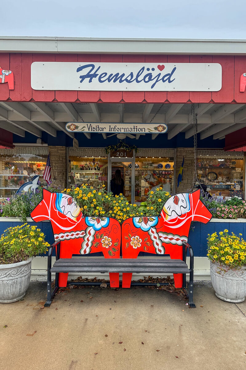 A bench with 2 red Dala horses painted on it in front of a store