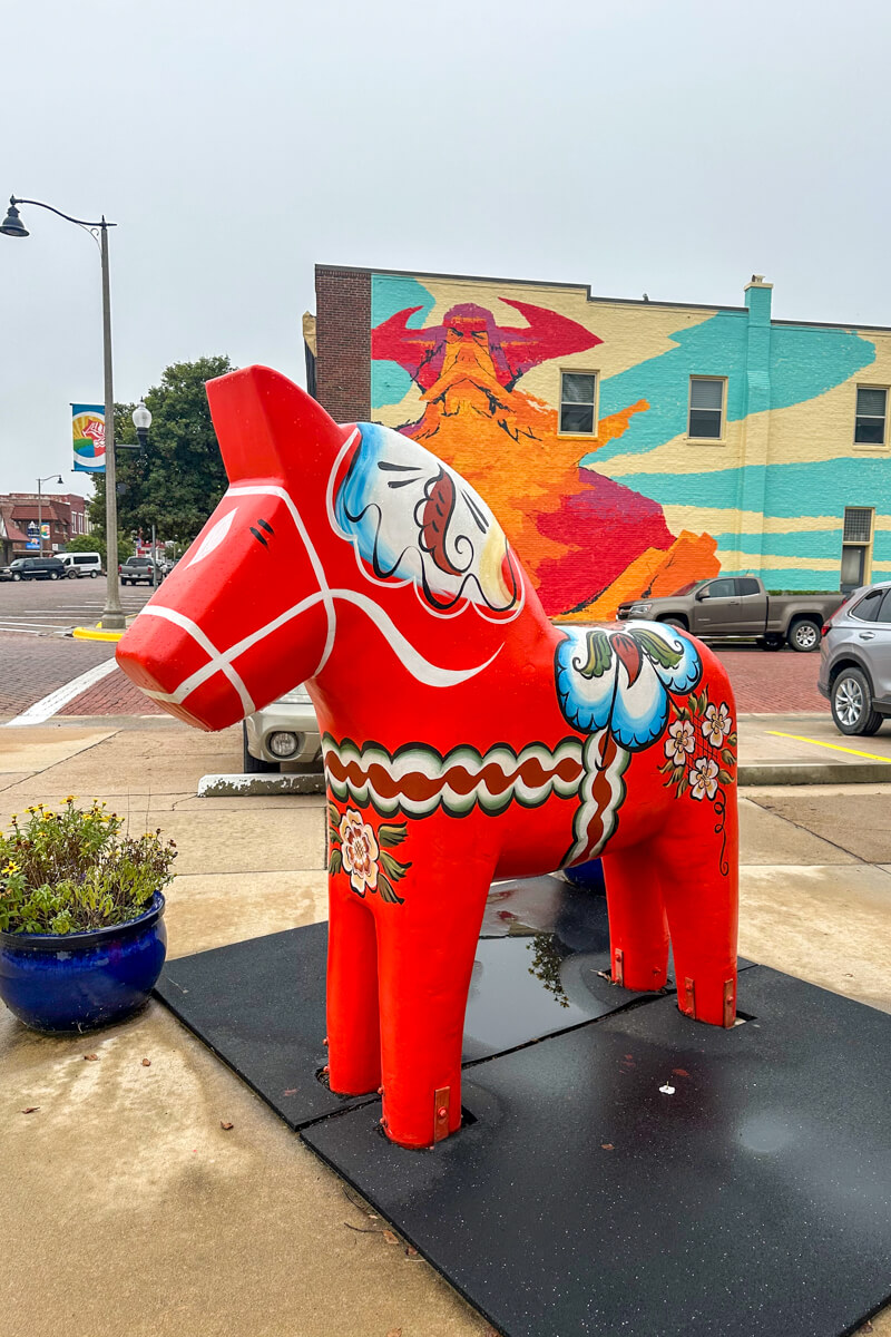 A classic red Dala Horse, pony sized, in front of a building with a Viking mural