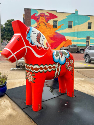 A classic red Dala Horse, pony sized, in front of a building with a Viking mural