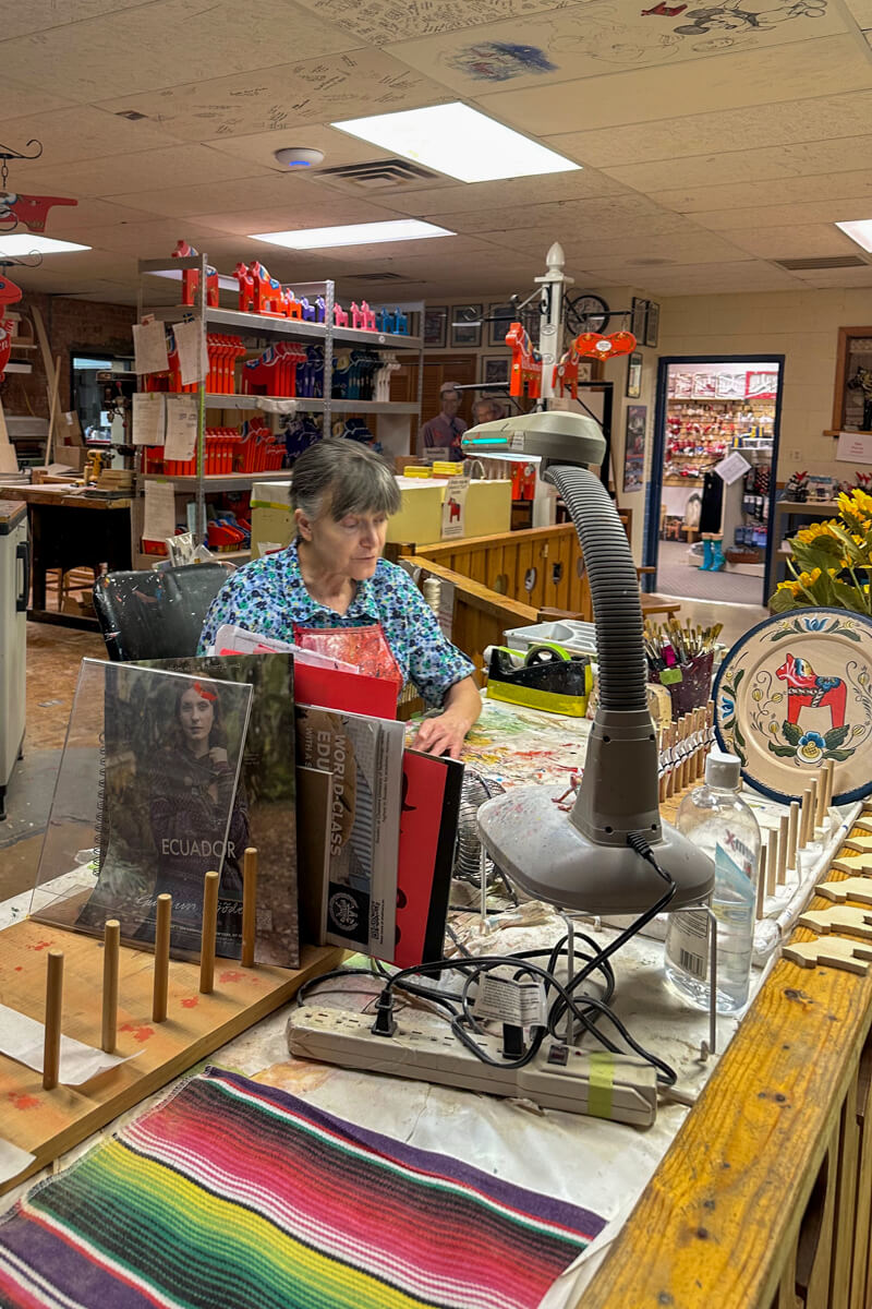 A gray haired woman sitting at her work table in the workshop with paint supplies spread around