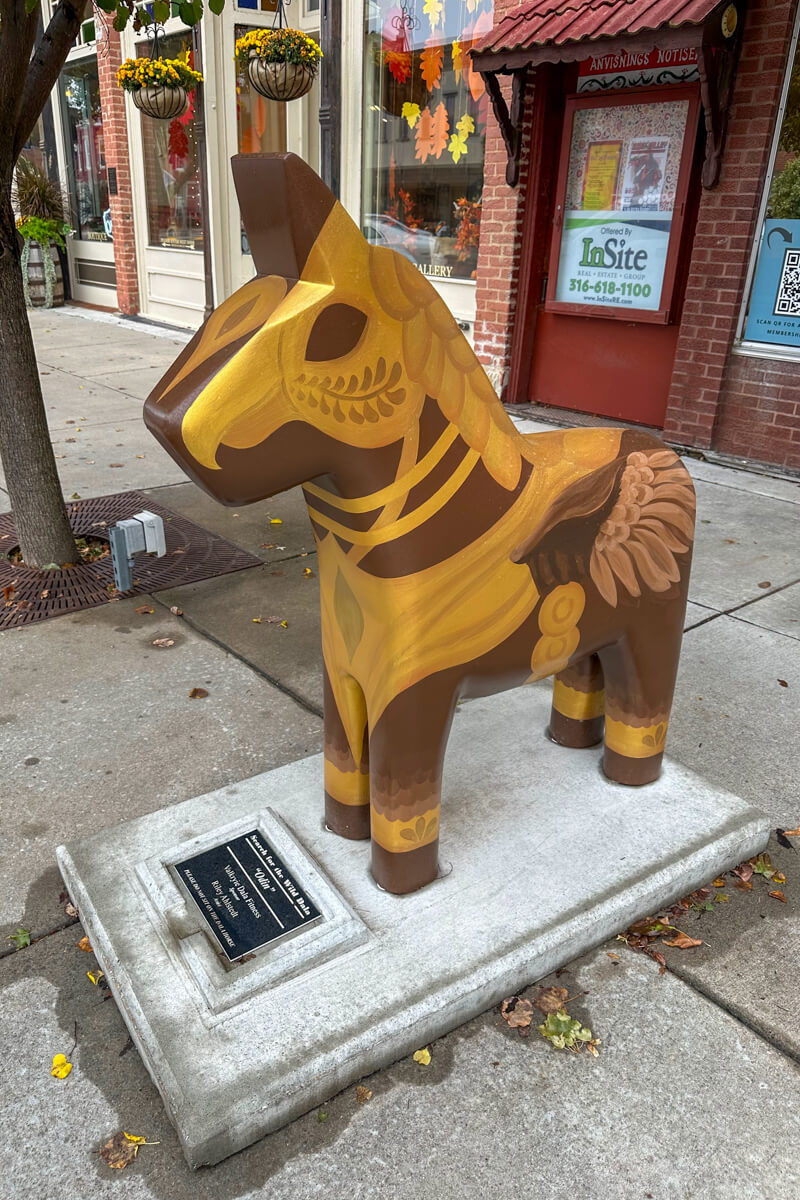 A Dala Horse, painted brown with gold decoration, pony sized, on the sidewalk in front of a store