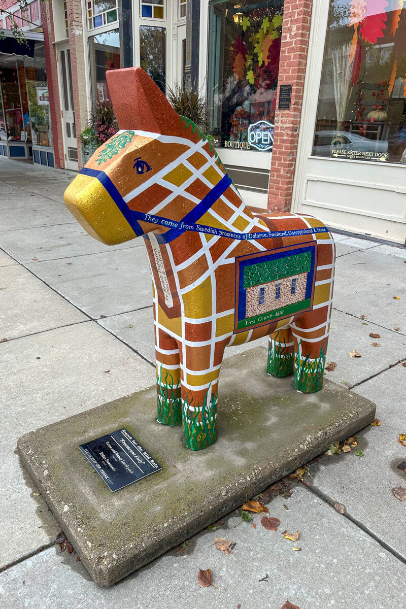 A Dala Horse, with a brown and gold street map painted on the side, pony sized, on the sidewalk in front of a store
