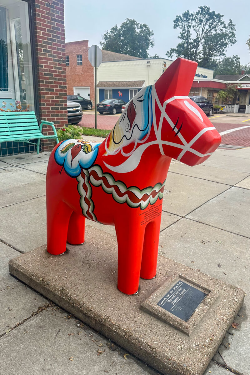 A classic red Dala Horse, pony sized, on the sidewalk in front of a store