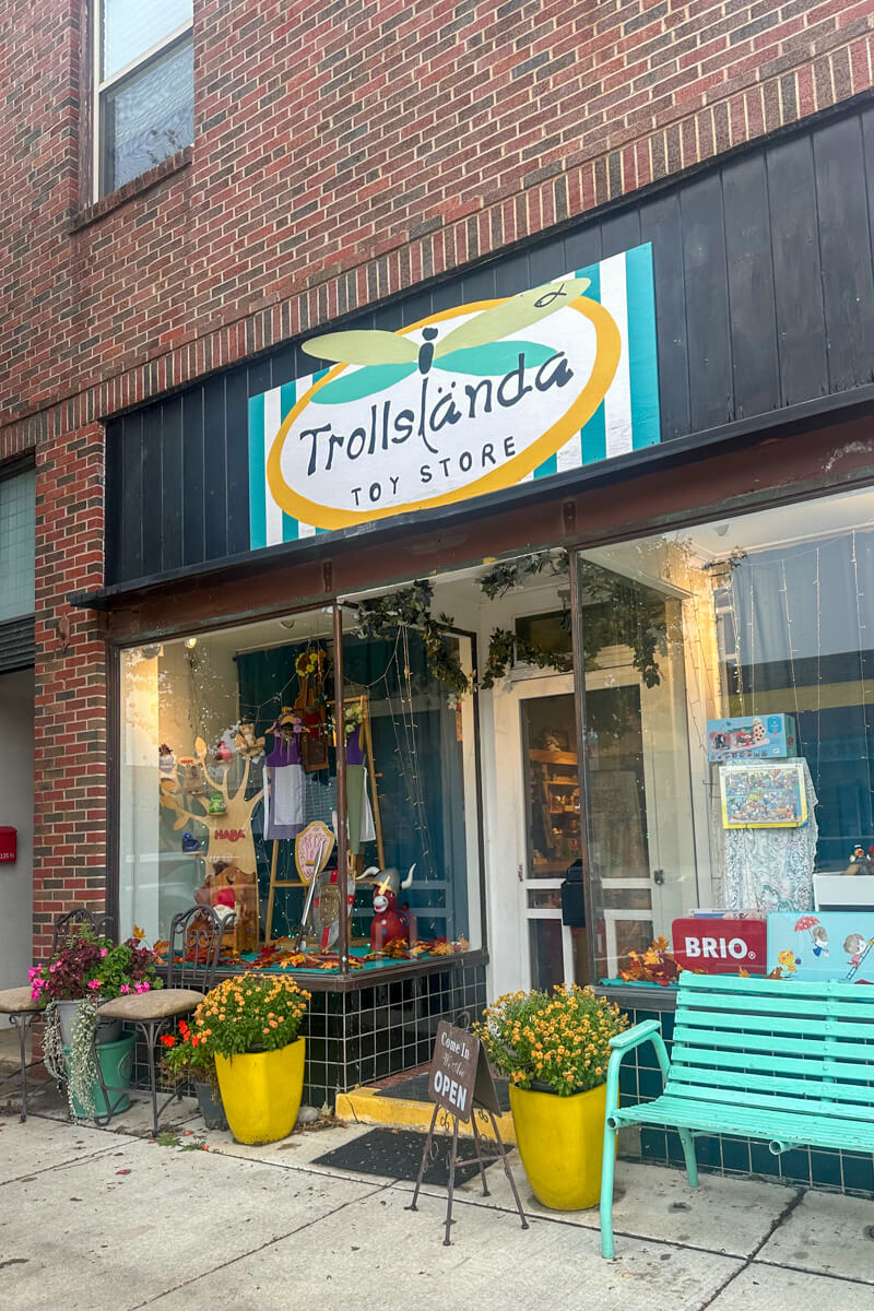 A store front of a toy shop with a teal bench and two yellow planters filled with mums