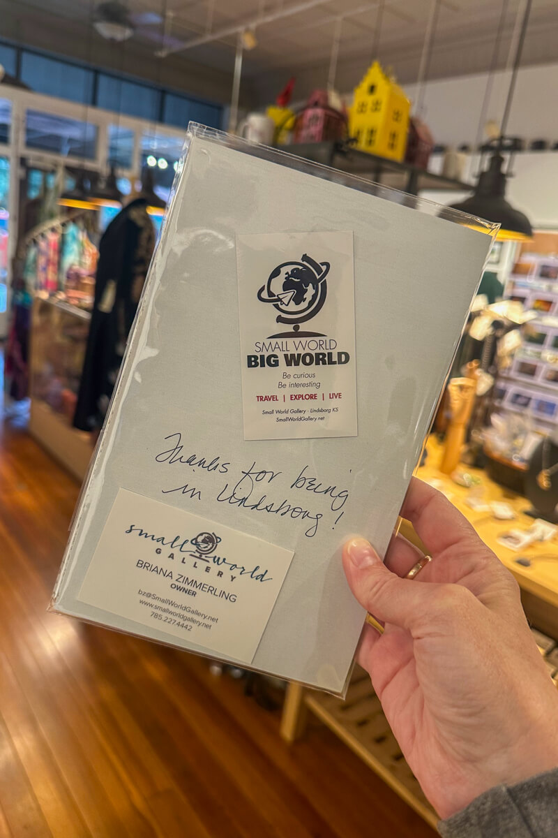 A hand holding up a white card with writing on it in front of the inside of the Small World Gallery
