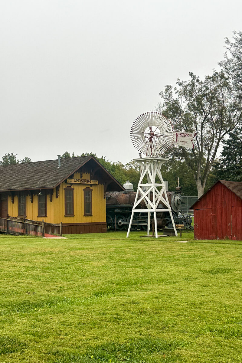 A grassy area in front of a traditional windmill, train station building and small red barn