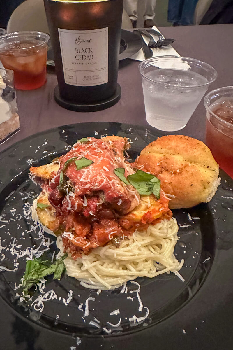 A black plate of gluten free pasta with meat and veggies on top cooked in a marinara sauce. With a gluten free roll and clear plastic cup of water