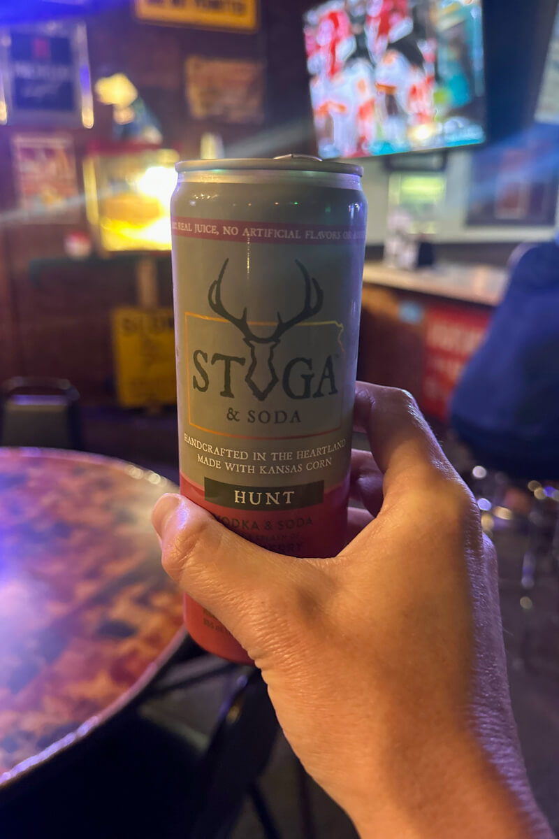 A hand holding up a can of Stuga seltzer with tables and tvs in the background