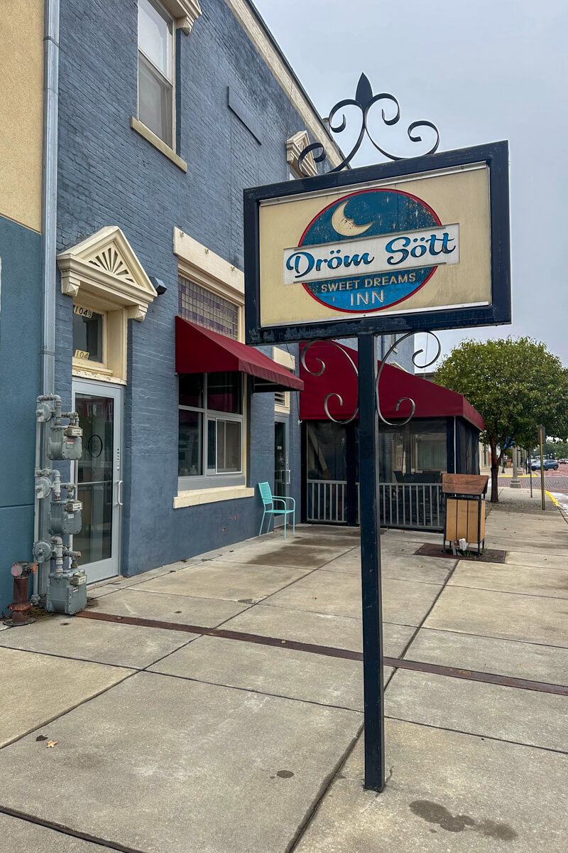 A blue and cream sign in a metal frame on the sidewalk in front of a painted blue brick building