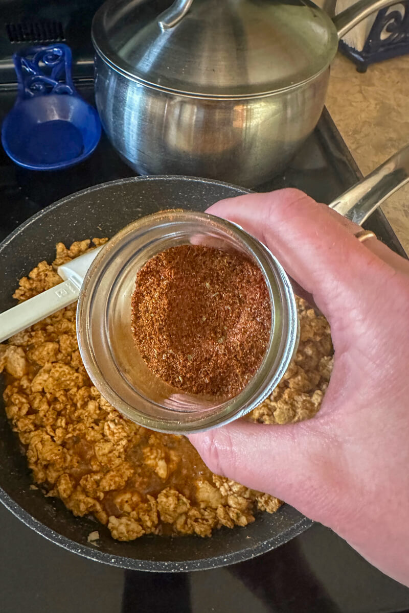 A hand holding a jar of chicken taco seasoning in front of a pan cooking ground chicken taco meat