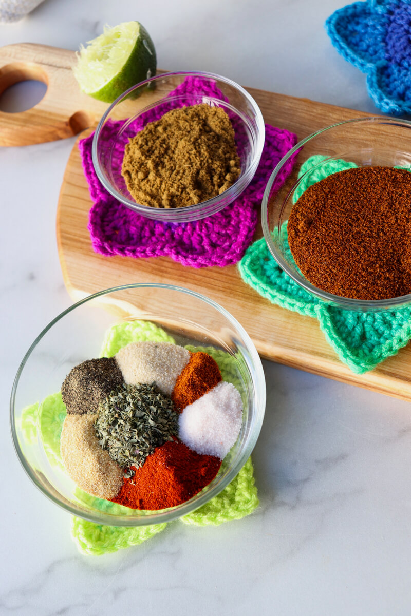 Overhead view of colorful spices in the chicken taco seasoning in a glass bowl with another smaller bowl and spice behind it on a wood cutting board crocheted star coaster
