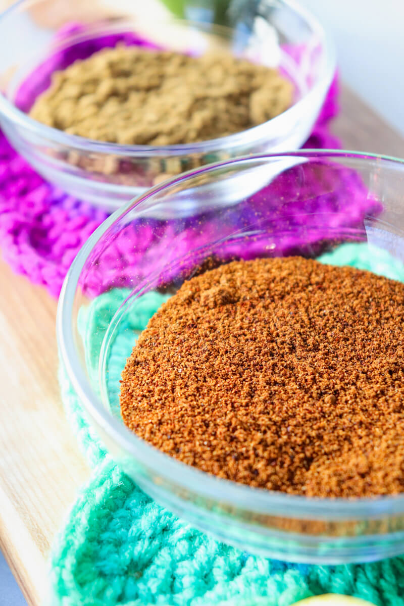 A close up of a dark orange spice, smoked paprika, in a small glass bowl in front of a smaller glass bowl of cumin