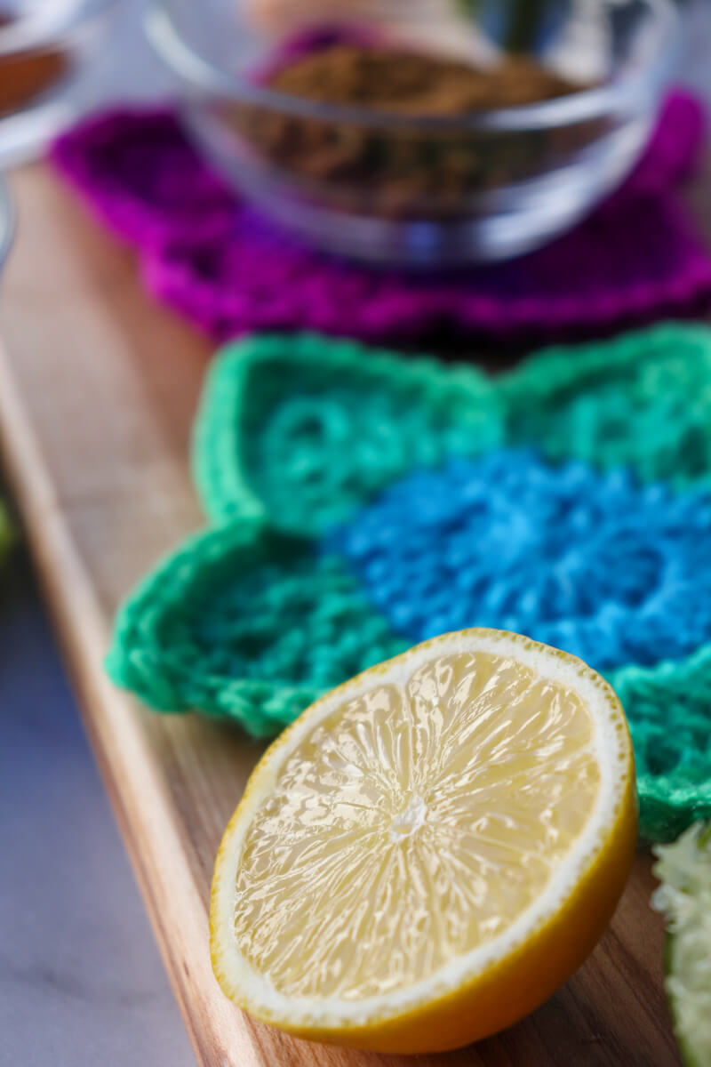 A half a lemon on a wooden cutting board in front of a green crocheted coaster, bhind that a magenta one with a glass bowl of spice