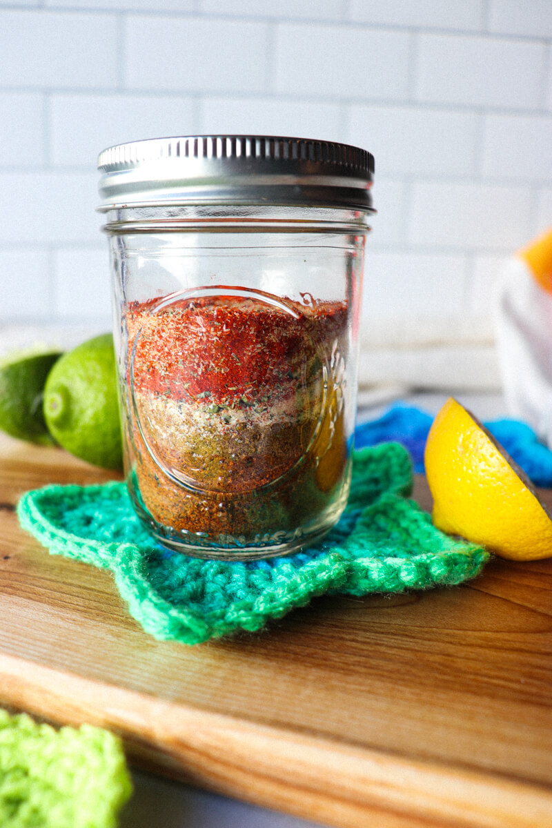 A glass Ball jar filled with layers of the spices in keto chicken taco seasoning on a crocheted green star coaster next to a lime and half a lemon