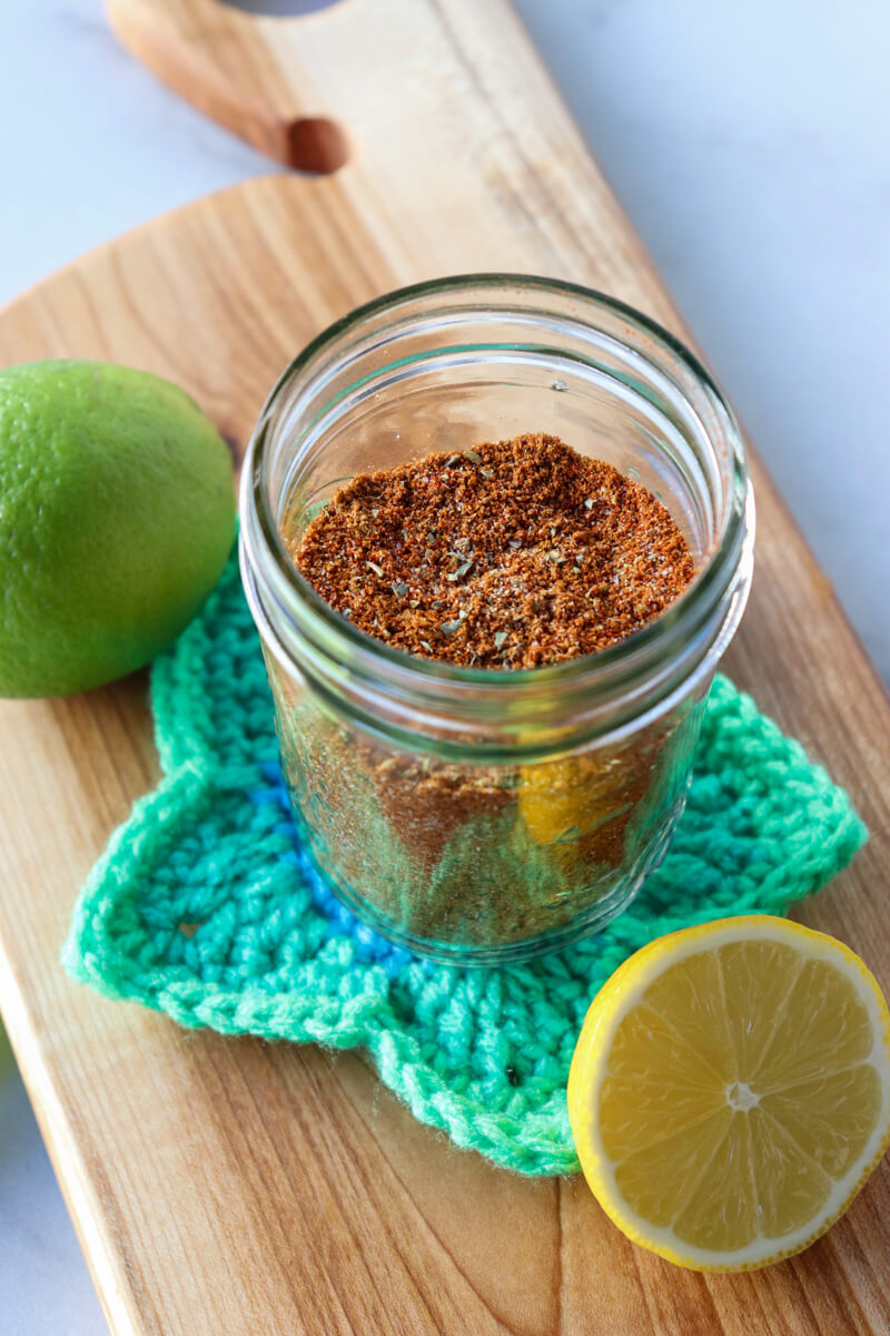 Front overhead view of a glass jar of easy chicken taco seasoning on a crocheted green star coaster next to a lime and half a lemon on a wooden cutting board