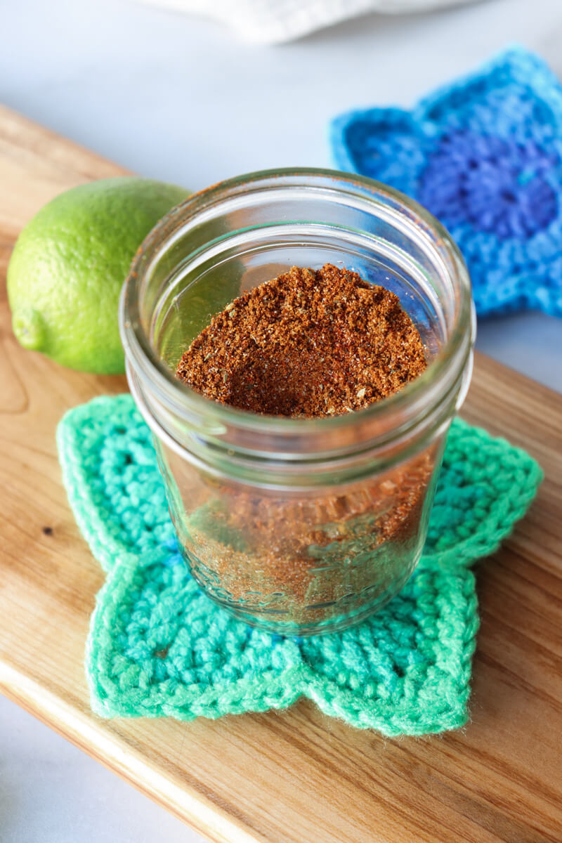 Front overhead view of a glass jar of easy chicken taco seasoning on a crocheted green star coaster next to a lime and half a lemon on a wooden cutting board with a blue star coaster in the background