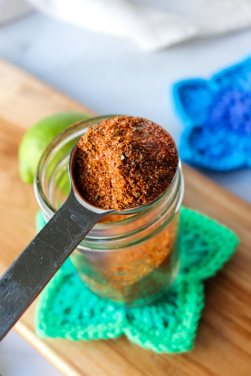 Front overhead view of a metal spoon of chicken taco seasoning in front of a glass jar of it on a crocheted green star coaster