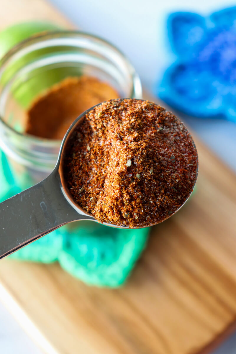 Front overhead view of a metal spoon of chicken taco seasoning in front of a glass jar of it on a crocheted green star coaster