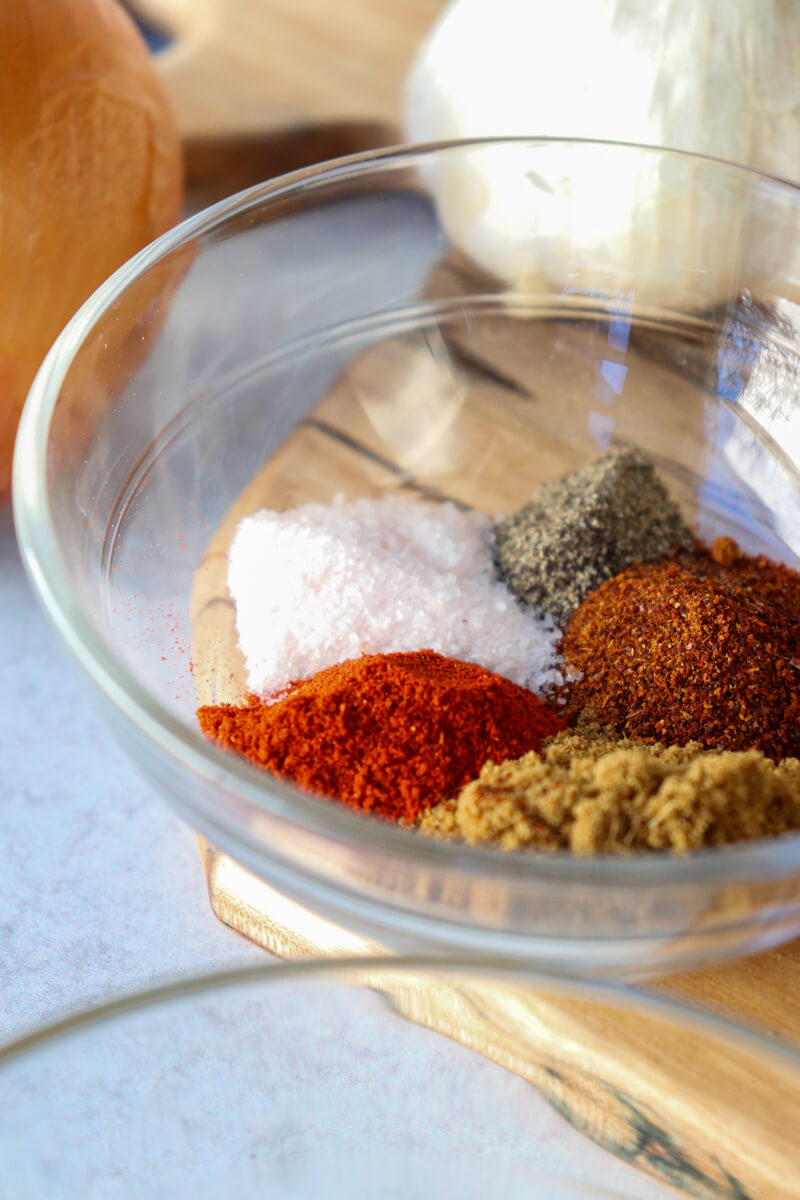 A clear glass bowl of spices in between an onion and garlic bulb