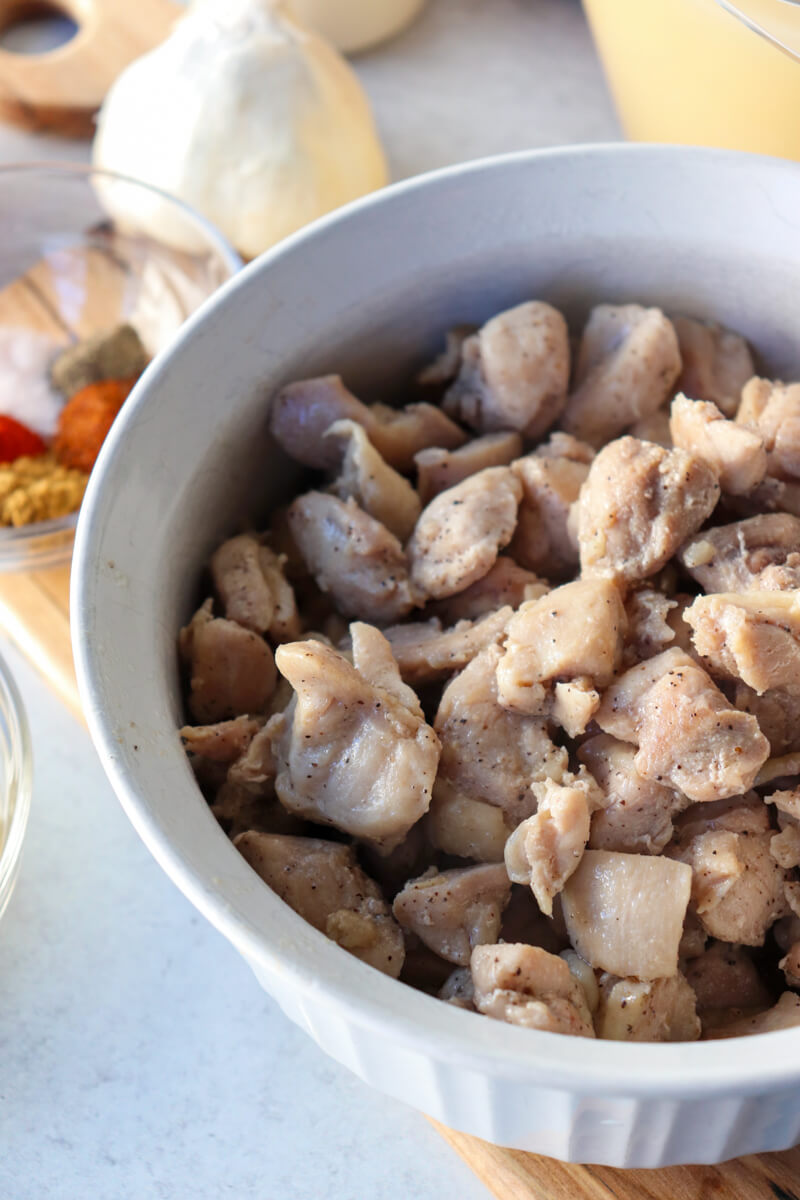 Overhead view of a white pyrex bowl of cooked chicken pieces on a wooden cutting board with garlic in the background.