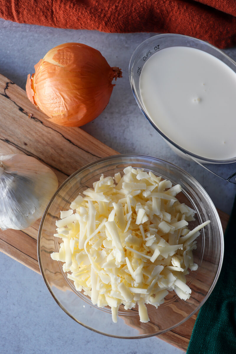 An overhead view of clear glass bowl of shredded white cheese on a wooden cutting board, a measuring cup of heavy cream, with an onion and garlic bulb.