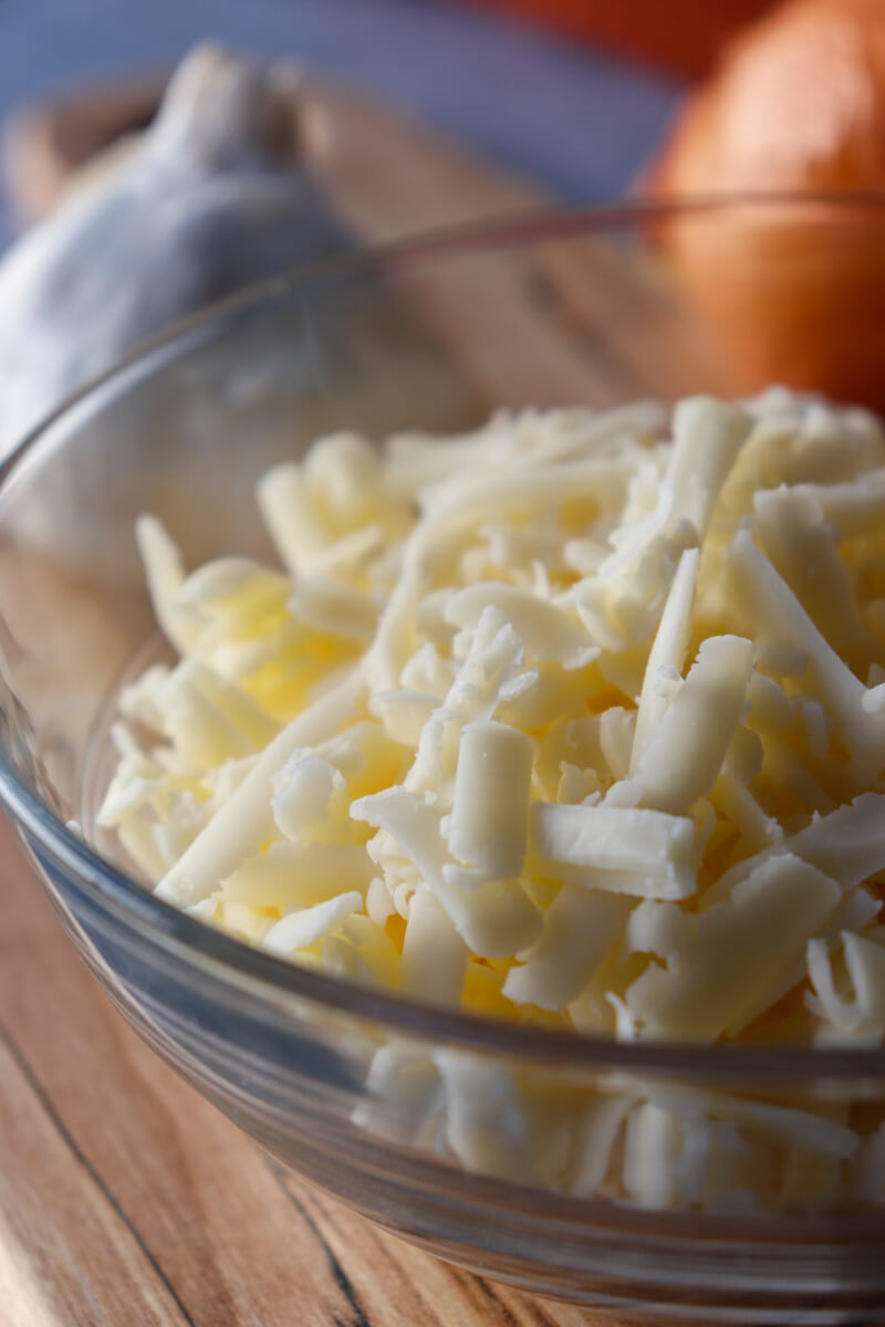 A clear glass bowl of shredded white cheese on a wooden cutting board with an onion and garlic bulb.