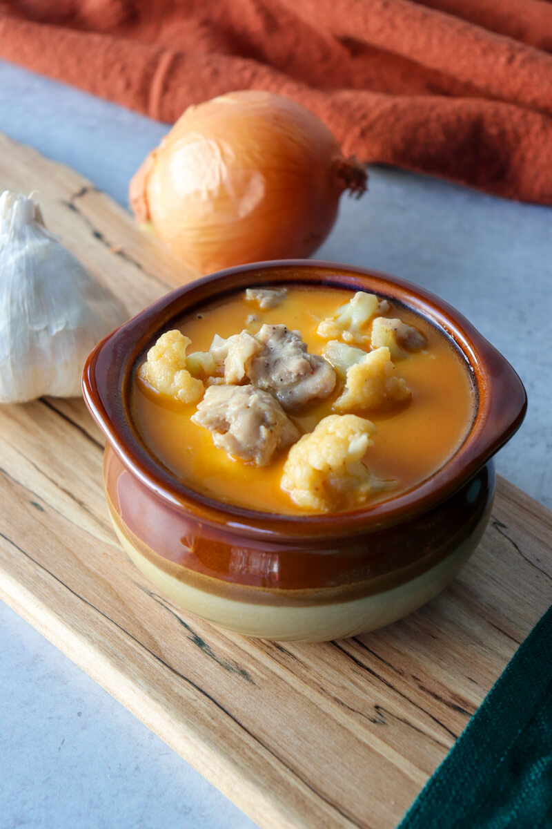 Overhead view of a brown ceramic bowl of keto white chicken chili soup with an onion behind it. On a wooden cutting board with a garlic bulb.