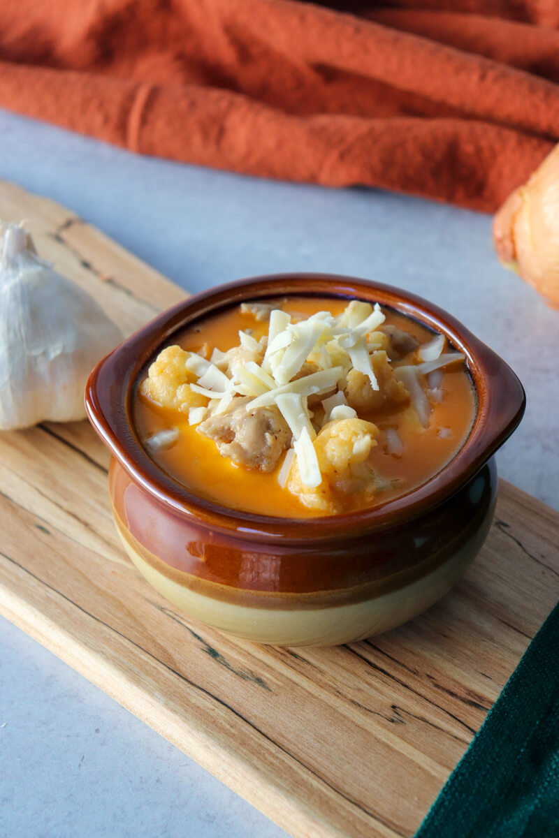 Overhead view of a brown ceramic bowl of keto white chicken chili soup with shredded Jack cheese. On a wooden cutting board with a garlic bulb.