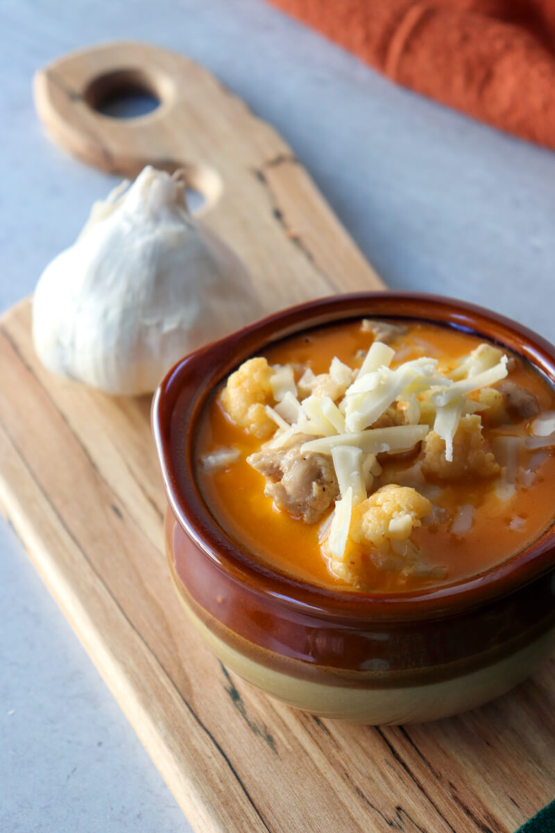Overhead view of a brown ceramic bowl of keto white chicken chili soup with shredded Jack cheese. On a wooden cutting board with a garlic bulb.