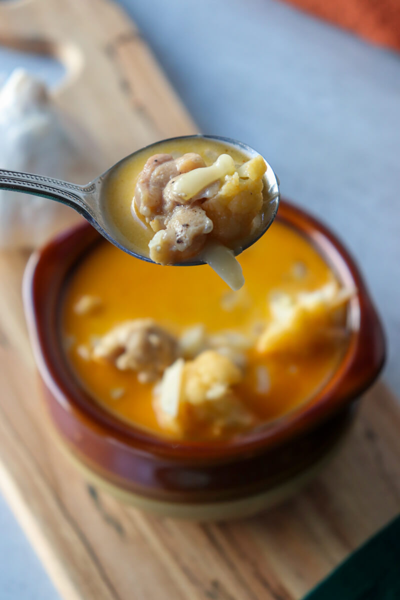 Overhead view of a brown ceramic bowl of keto white chicken chili soup with a spoon of it held up above it. On a wooden cutting board with a garlic bulb.