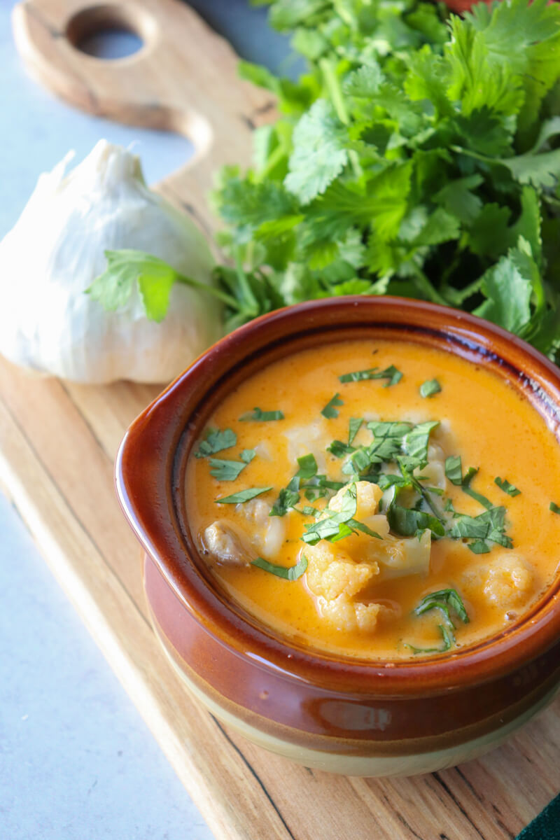 Overhead view of a brown ceramic bowl of keto white chicken chili with chopped cilantro, a bunch of cilantro and garlic in the background, on a wooden cutting board.