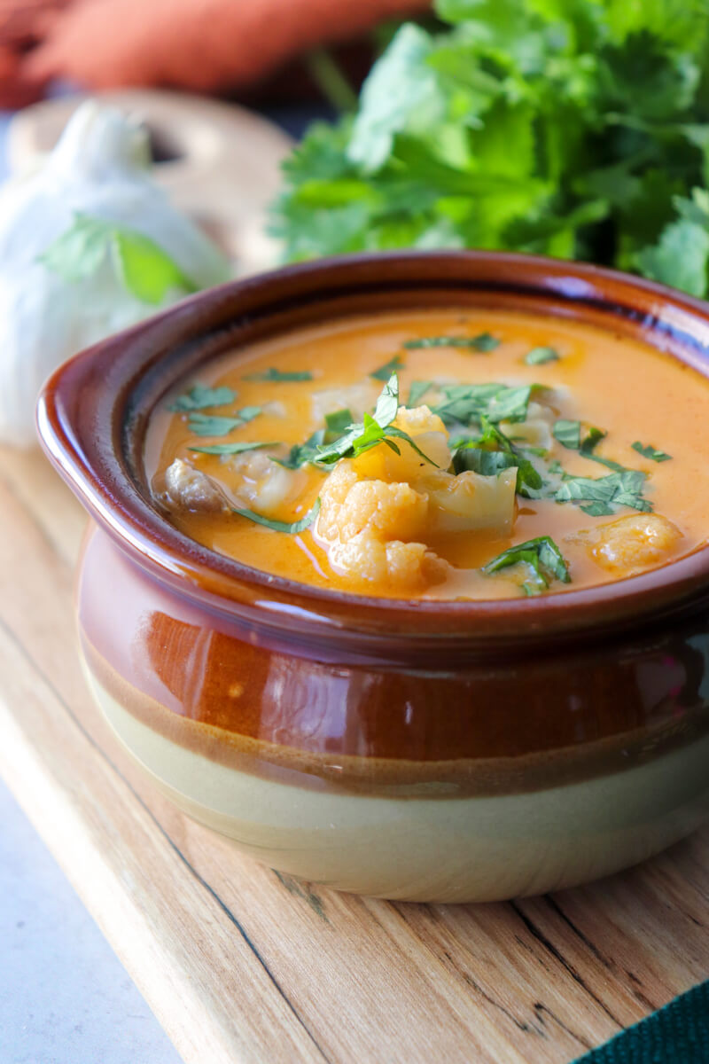 Front view of a brown ceramic bowl of keto white chicken chili with chopped cilantro, a bunch of cilantro and garlic in the background, on a wooden cutting board.