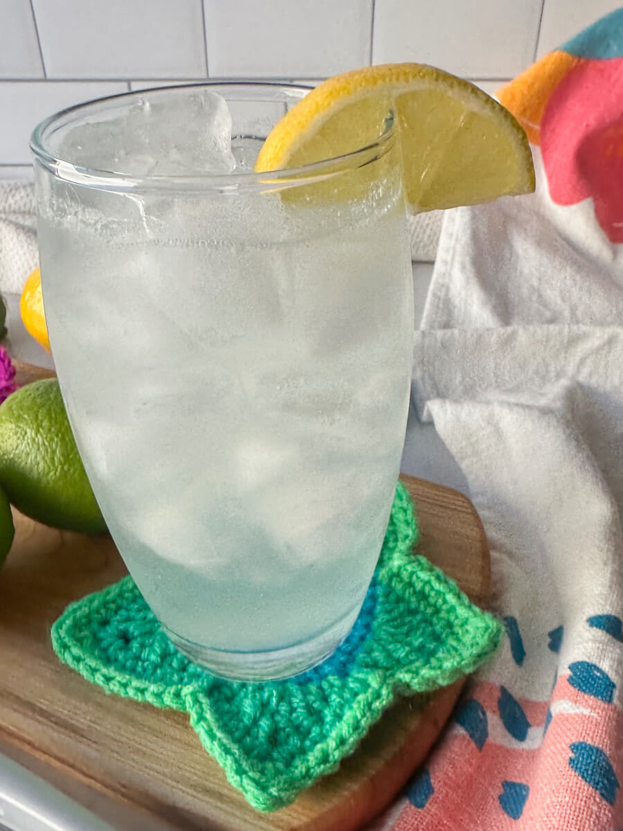 Closeup view of a clear glass filled with a low carb electrolyte drink, pale color, with ice and a lemon on the rim of the glass. Sitting on a wooden cutting board and crocheted green star coaster.